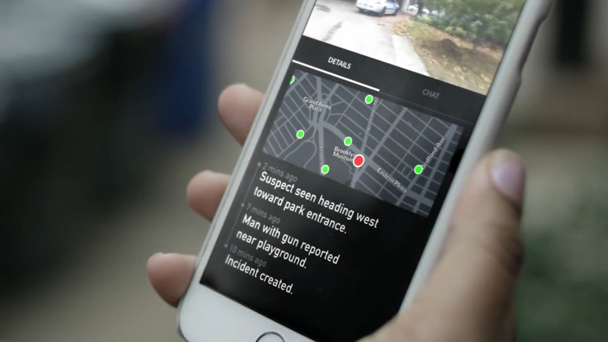 A person's hand holding a phone with the Citizen app open. The app shows a map and text that says "Suspect seen heading west toward park entrance."