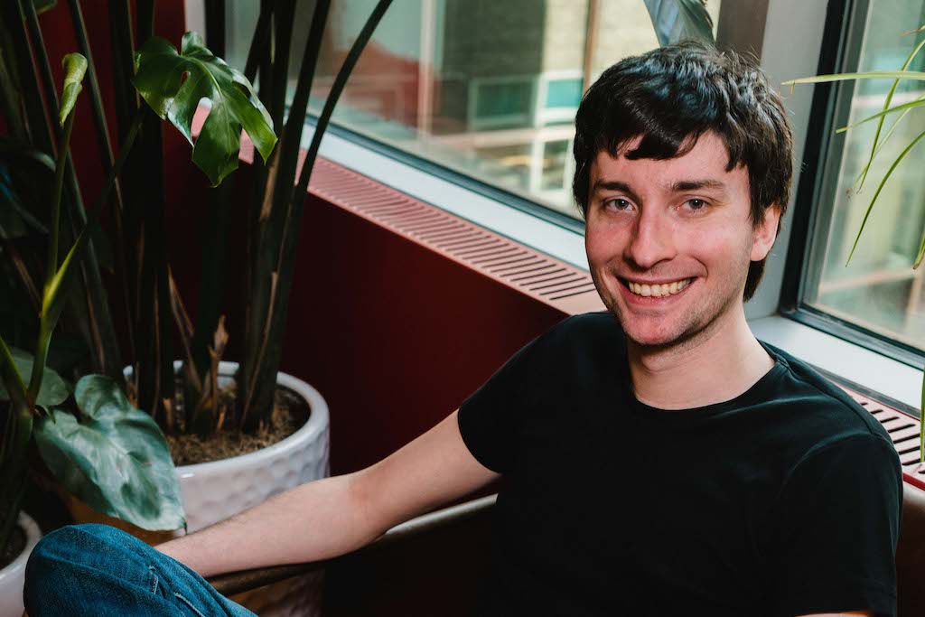 A photo of Andrew Harrison sitting on a chair, with a red wall and plants in the background.
